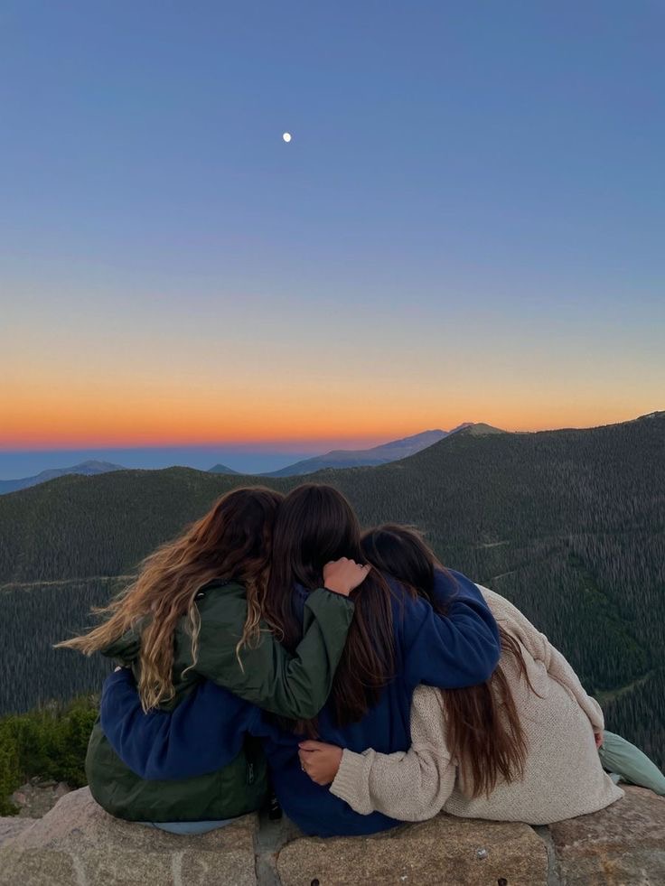 Três mulheres sentadas juntas diante de uma paisagem montanhosa ao entardecer, abraçadas enquanto observam o horizonte. O céu apresenta tons de azul, laranja e rosa, com a lua visível ao alto.